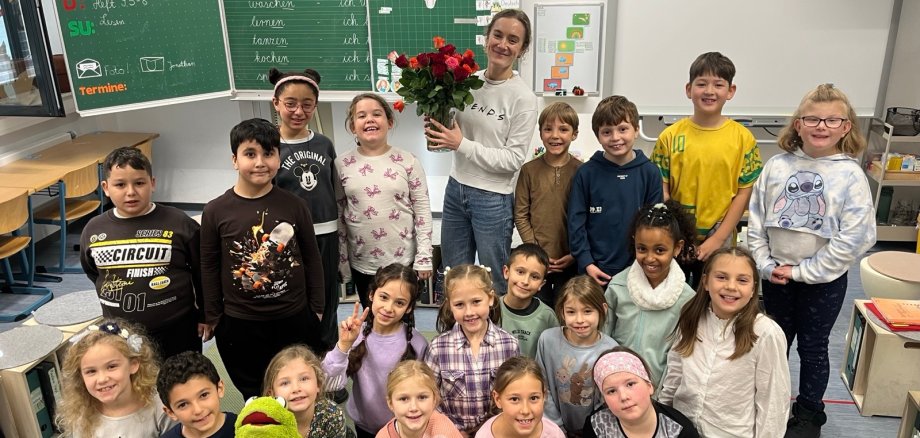 Herzlichen Glückwunsch, Frau Scheffel! Frau Scheffel hält einen Strauß Rosen in der Hand, den sie von allen SchülerInnen der Klasse 2a überreicht bekommen hat. Alle Kinder der Klasse 2a sitzen und stehen um sie herum und freuen sich!