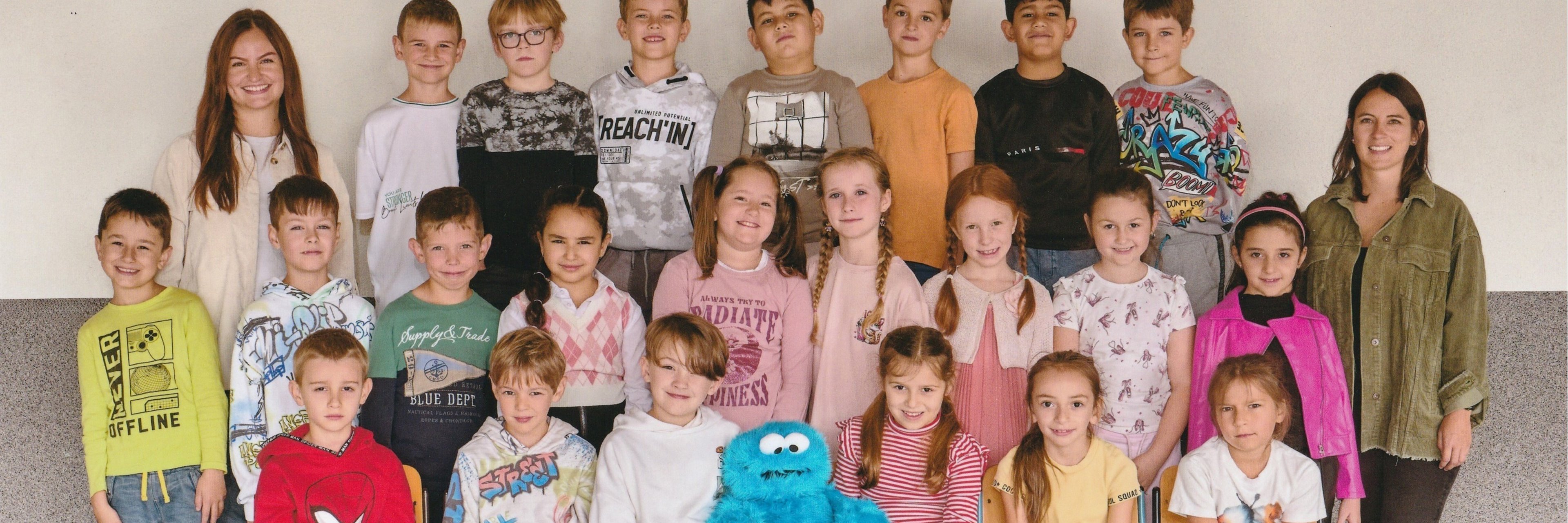 22 Kinder und 2 Lehrkräfte und das Krümelmonster der Klasse 2b stehen und sitzen für das Klassenfoto bereit.