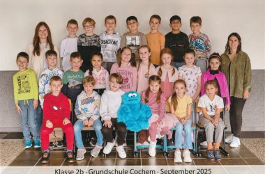 22 Kinder und 2 Lehrkräfte und das Krümelmonster der Klasse 2b stehen und sitzen für das Klassenfoto bereit.